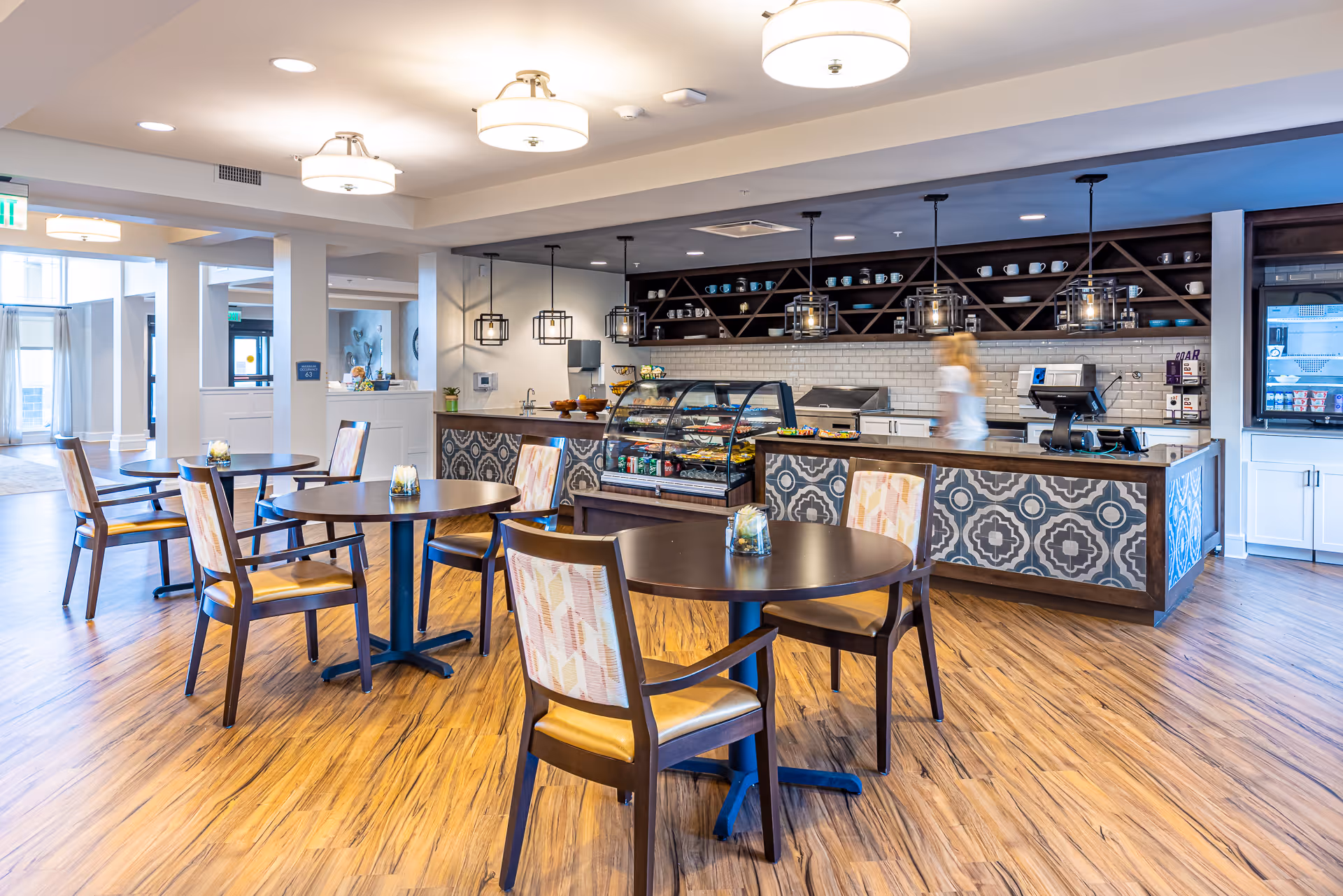 A bright and modern dining area in a senior living facility featuring round wooden tables with patterned cushioned chairs. The room has wood flooring and a counter area with a display case containing food items. Pendant lights hang above the counter, and shelves with cups and bowls are visible behind it. A person is walking behind the counter, and large windows allow natural light to fill the space.