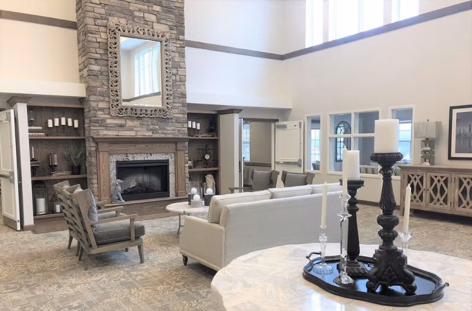 A spacious senior living facility common area with a stone fireplace, a large ornate mirror above it, and built-in shelves on either side. The room features comfortable seating including a beige sofa and gray armchairs, a patterned carpet, and a marble table with decorative candle holders in the foreground. Large windows near the ceiling allow natural light to fill the room.