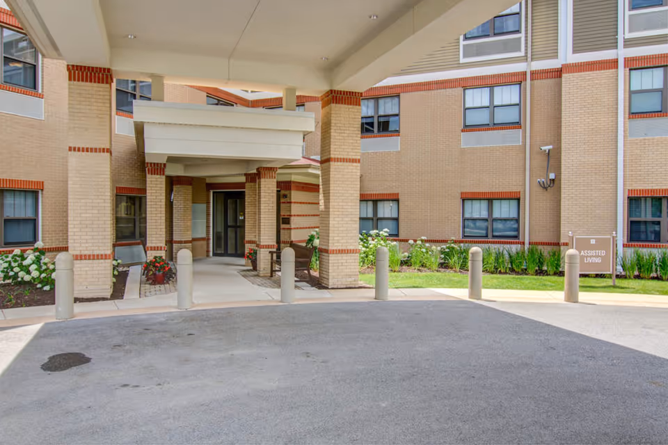 Entrance area of EverTrue Lutheran Hillside Village showing a covered drop-off zone with brick pillars and a bench. The building exterior is beige brick with red brick accents and multiple windows. There are plants and flowers along the building and a sign indicating 'Assisted Living'.