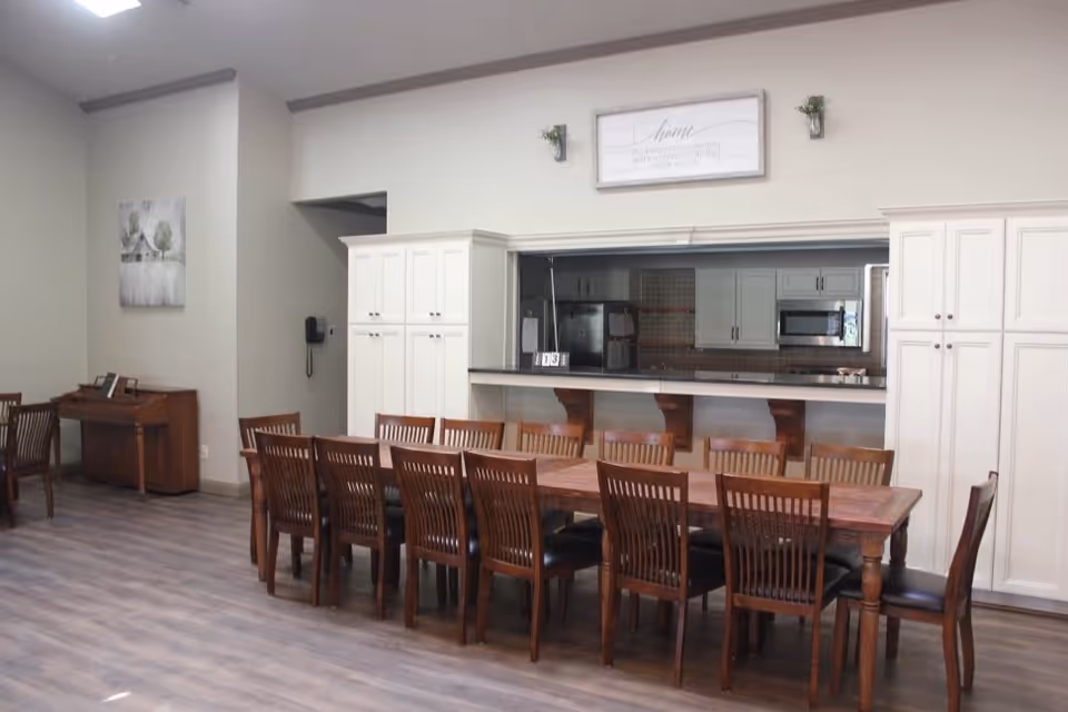 A spacious dining area with a long wooden table surrounded by multiple wooden chairs. In the background, there is a kitchen with white cabinets, a refrigerator, microwave, and a pass-through counter with wooden supports. On the left side, there is a piano against the wall with a framed picture above it. The flooring is light-colored wood, and the walls are painted a light neutral color. A decorative sign and small plants are mounted above the kitchen pass-through window.