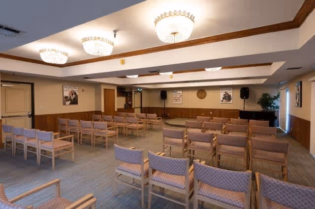 A large room with multiple rows of wooden chairs with padded seats and backs arranged facing a small stage area with two speakers and a piano. The room has beige walls with wooden paneling on the lower half, three chandeliers on the ceiling, and framed pictures on the walls.
