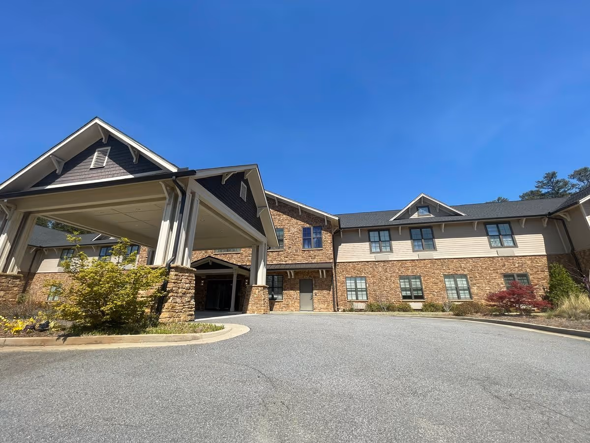 Front entrance of a two-story brick-faced senior living building with a covered porte-cochère under a clear blue sky.