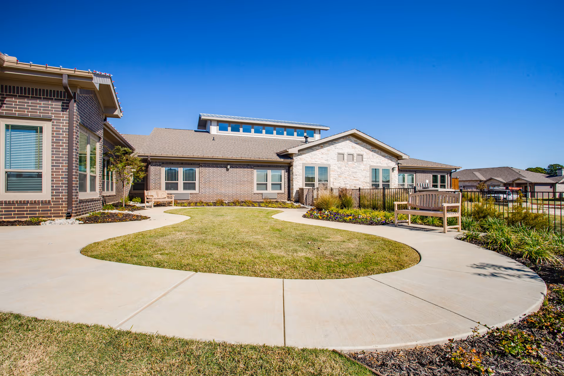 Outdoor courtyard area of a senior living facility with a circular concrete walkway surrounding a grassy lawn. There are wooden benches along the walkway, flower beds, and a brick and stone building in the background under a clear blue sky.