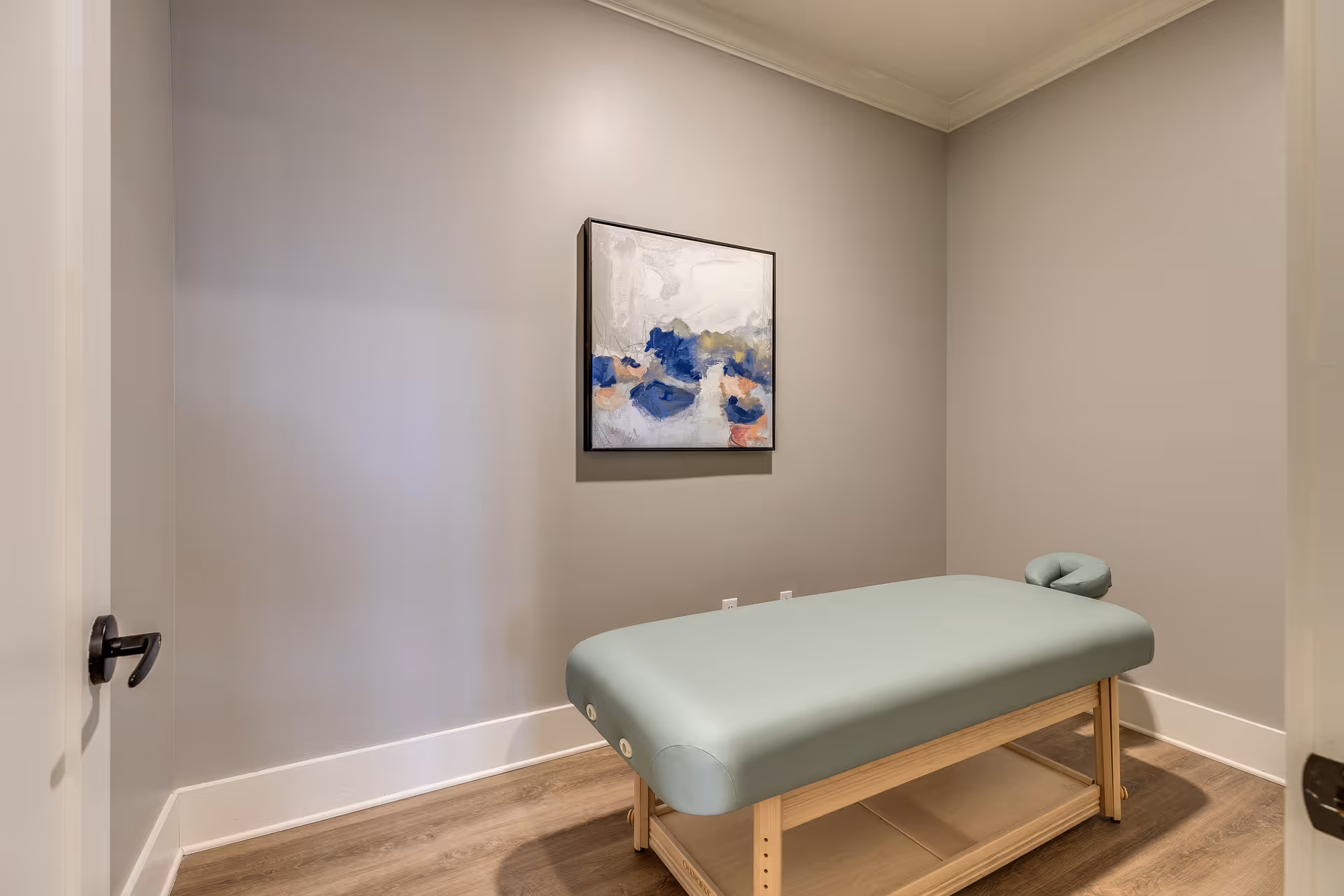 A small room with light gray walls featuring a massage table with a light green cushion and wooden legs. A modern abstract painting with blue, white, and peach colors hangs on the wall above the table. The floor is wood, and the door with a black handle is partially open.