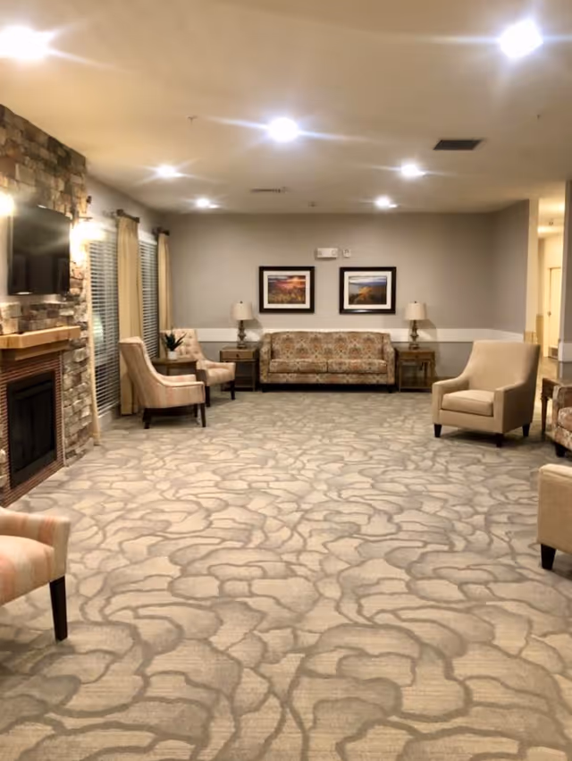 Spacious senior living lounge with patterned carpet, sofas and armchairs, a stone fireplace with mounted TV, and framed art on the far wall.