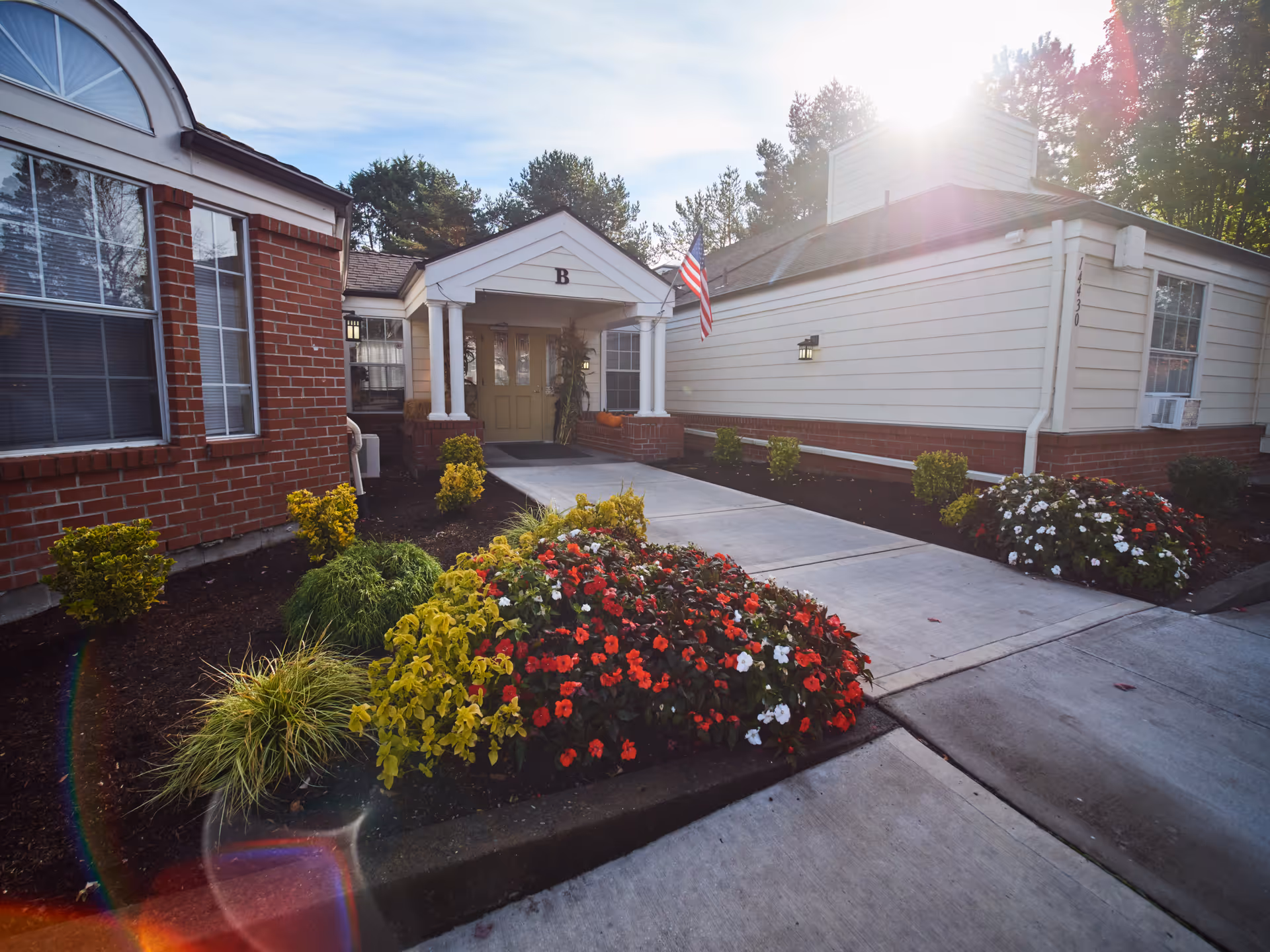 Exterior view of a senior living facility entrance with a concrete walkway leading to a door under a small portico labeled with the letter B. The building has brick and white siding walls, with an American flag mounted on the right side. There are landscaped flower beds with red, white, and yellow flowers and green shrubs on either side of the walkway. Sunlight is visible in the background through trees.