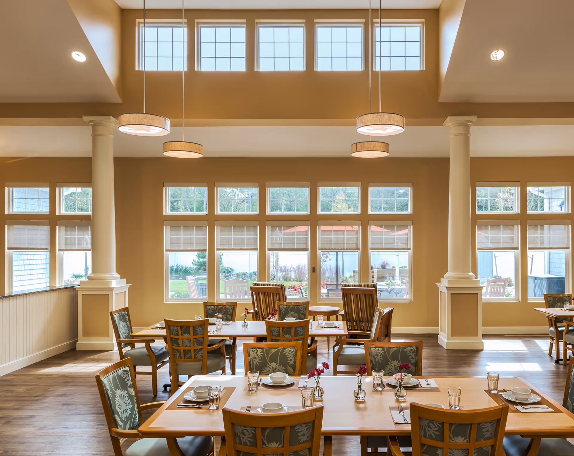 A bright dining room with large windows allowing natural light to fill the space. The room features wooden tables set with plates, glasses, and small vases with flowers. Comfortable chairs with patterned upholstery surround the tables. The walls are painted a warm beige color, and there are two white columns supporting the ceiling. Modern pendant lights hang from the ceiling, and outside the windows, a garden area with patio furniture and umbrellas is visible.