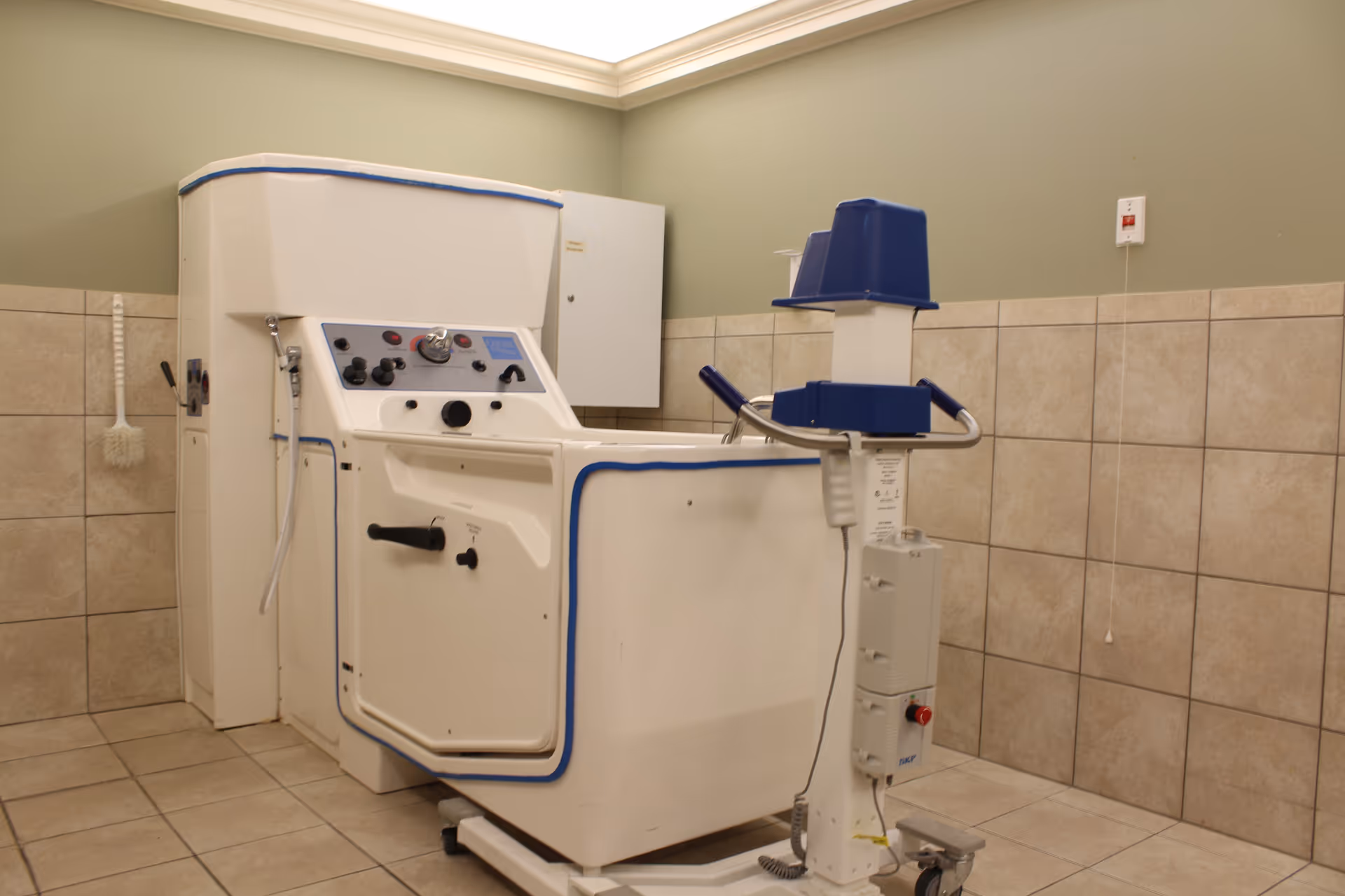 A specialized bathing tub with control knobs and a door for assisted bathing in a tiled room with light green walls, a white brush hanging on the wall, and a control panel on a stand with wheels.