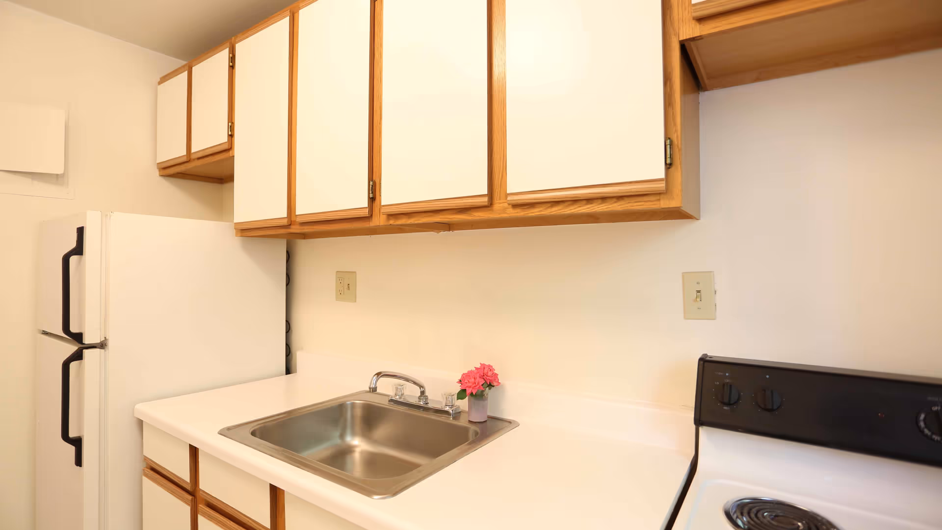 A small kitchen area with white countertops, a stainless steel sink, a white refrigerator with black handles, white cabinets with wooden trim, and a white electric stove with black control knobs. A small vase with pink flowers is placed next to the sink.