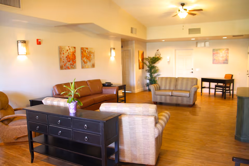 Bright communal living room with sofas, armchairs, a console table with a potted plant, wall art, and wood flooring.