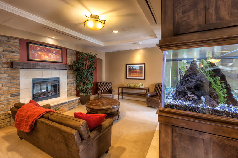 Cozy communal living room with a sofa and throw, fireplace, armchairs, round ottoman, and a large aquarium.