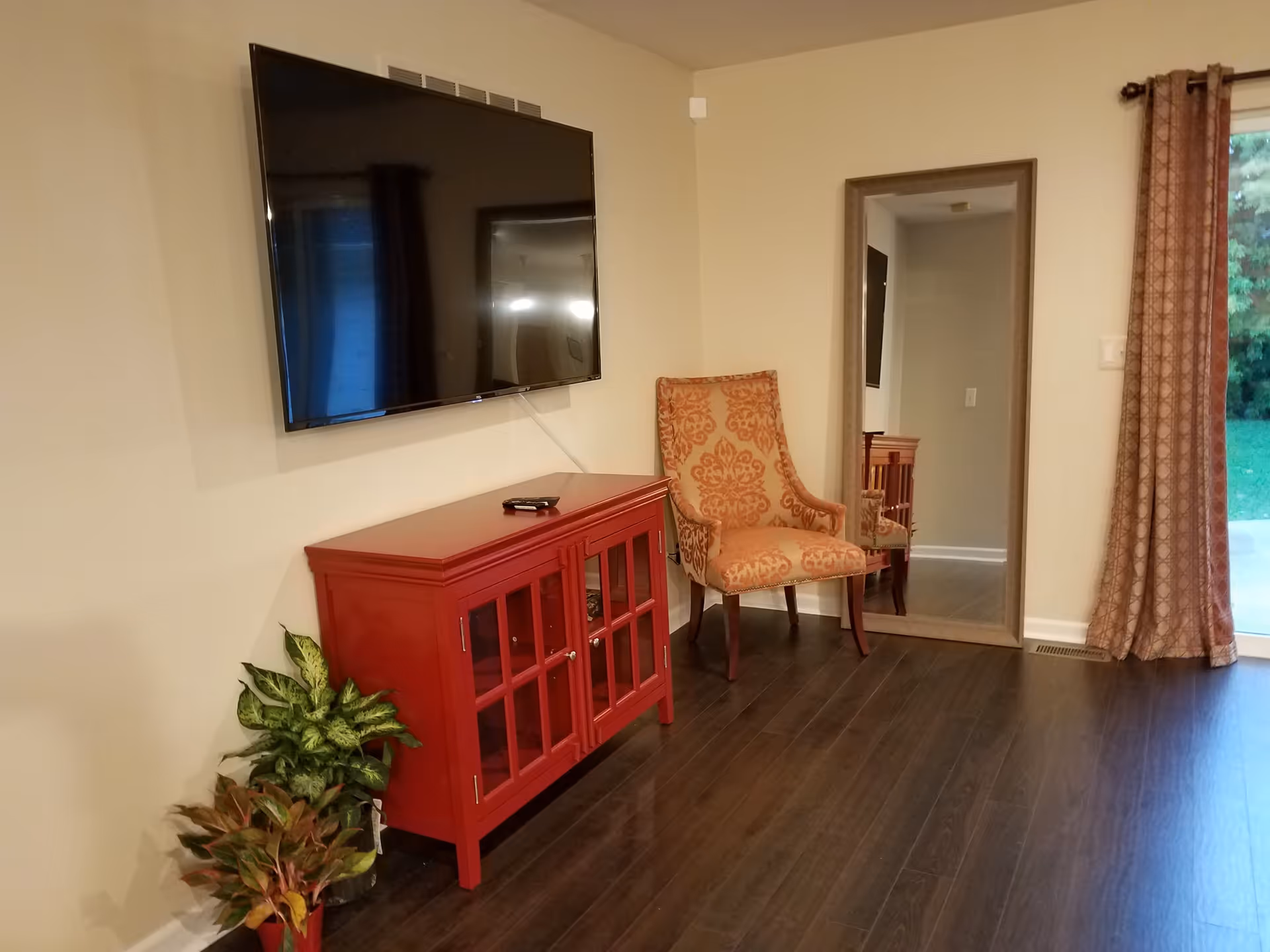 Living room with a wall-mounted TV above a red cabinet, a patterned armchair, a full-length mirror, potted plants, and a sliding glass door.
