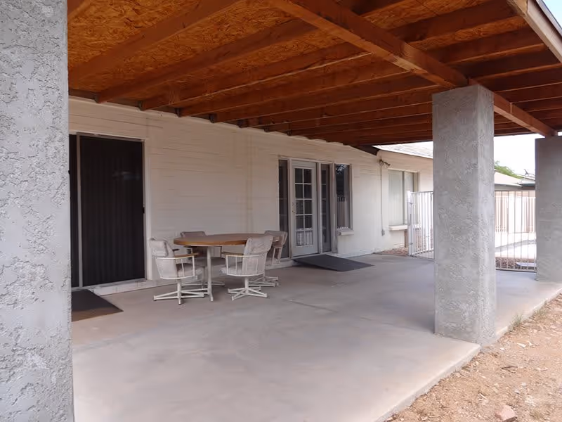 Covered outdoor patio area with a wooden ceiling, concrete floor, and two large supporting columns. There is a round table with four cushioned swivel chairs. The patio is adjacent to a white building with a black door, a set of French doors, and a window. A metal gate is visible on the right side.