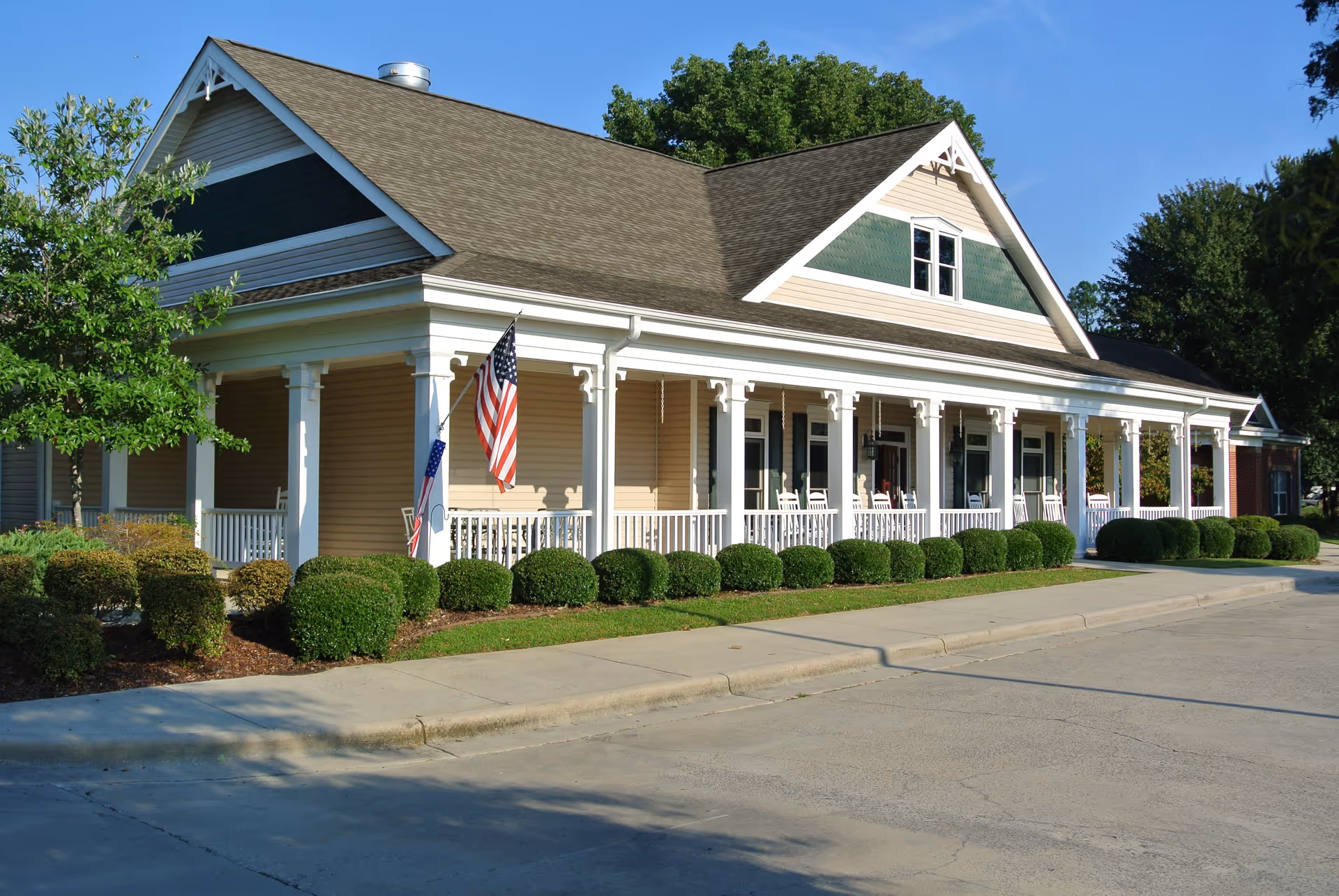 Exterior view of a single-story senior living facility building with a large covered porch featuring white columns and railings. The building has beige siding with green and cream accents near the roof. There are neatly trimmed bushes along the sidewalk and two American flags displayed on the porch. The sky is clear and blue.