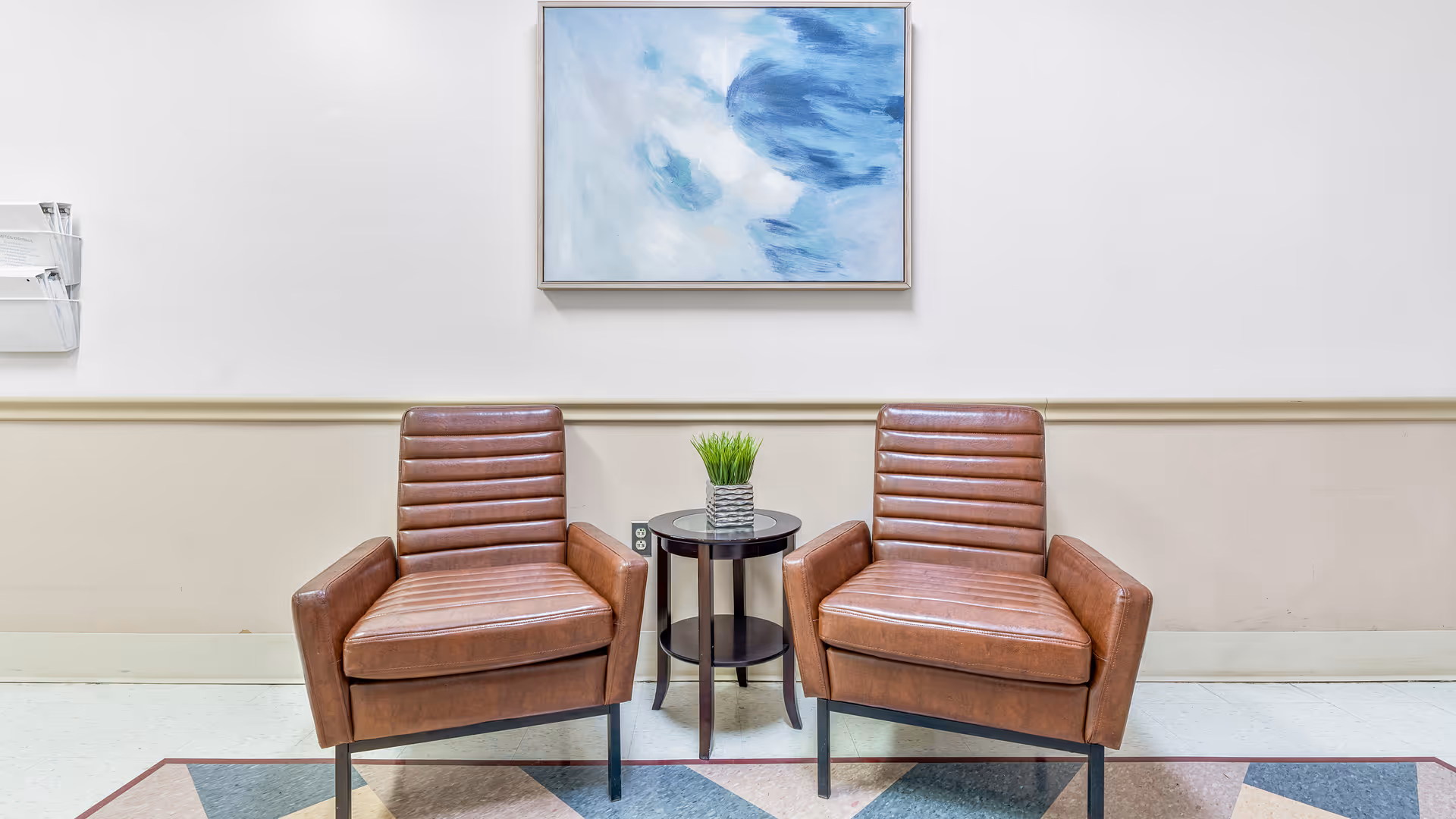 Two brown leather armchairs with horizontal stitching are placed side by side with a small round black table between them holding a small potted plant. Above the chairs hangs a framed abstract painting with blue and white colors. The setting is against a beige and white wall with a light-colored floor.