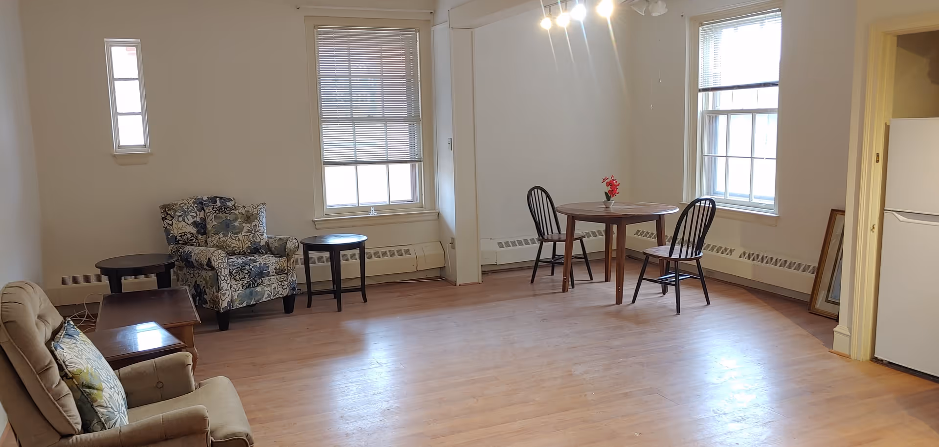 A bright room with wooden flooring featuring two armchairs, a small wooden table, a round dining table with two chairs, and two large windows with blinds. There is a white refrigerator in the corner and a mirror leaning against the wall.