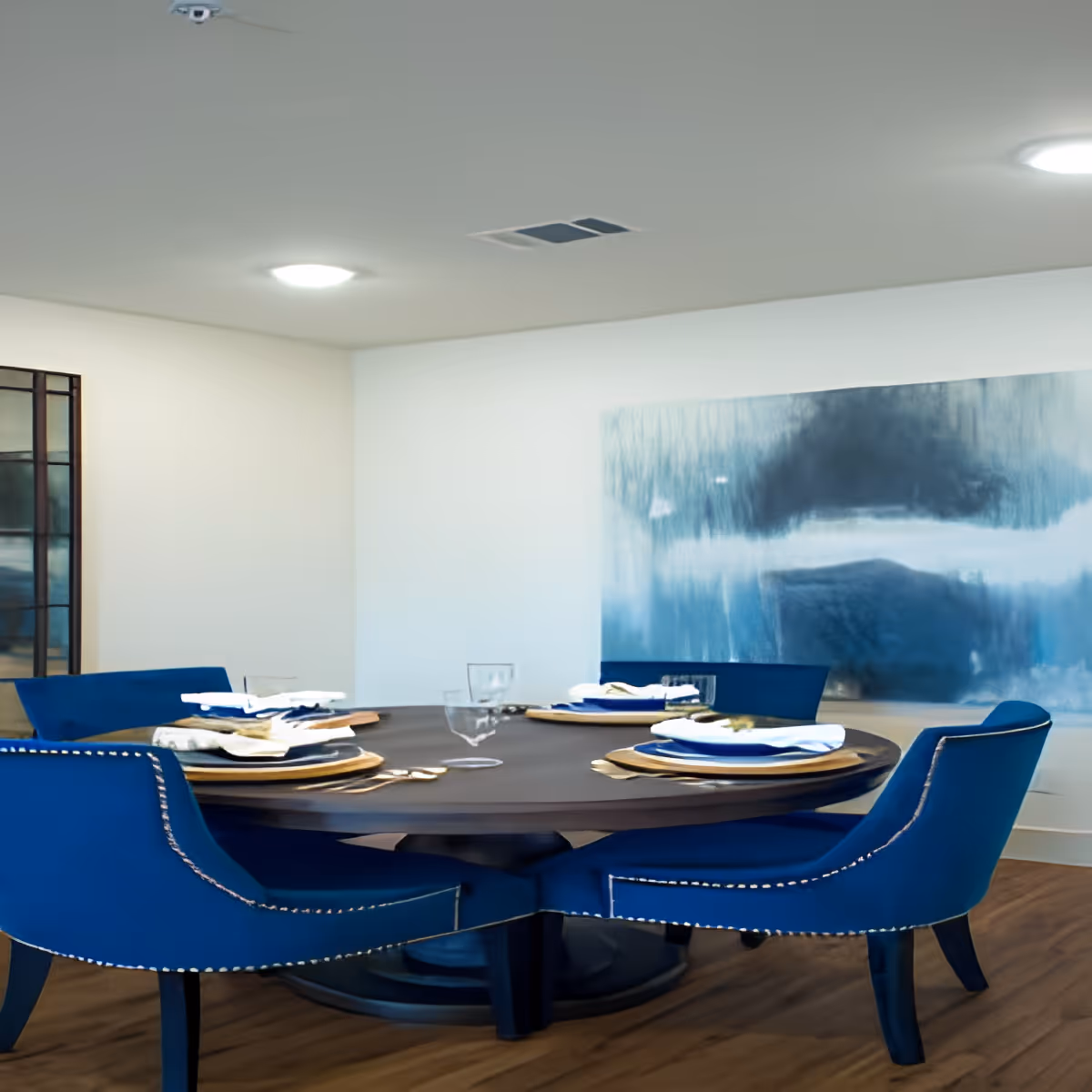 A modern dining room with a round dark wooden table set for four. The table is surrounded by four blue upholstered chairs with nailhead trim. The table is set with plates, napkins, glasses, and cutlery. The room has light-colored walls, a large abstract blue and gray painting on one wall, and wooden flooring.