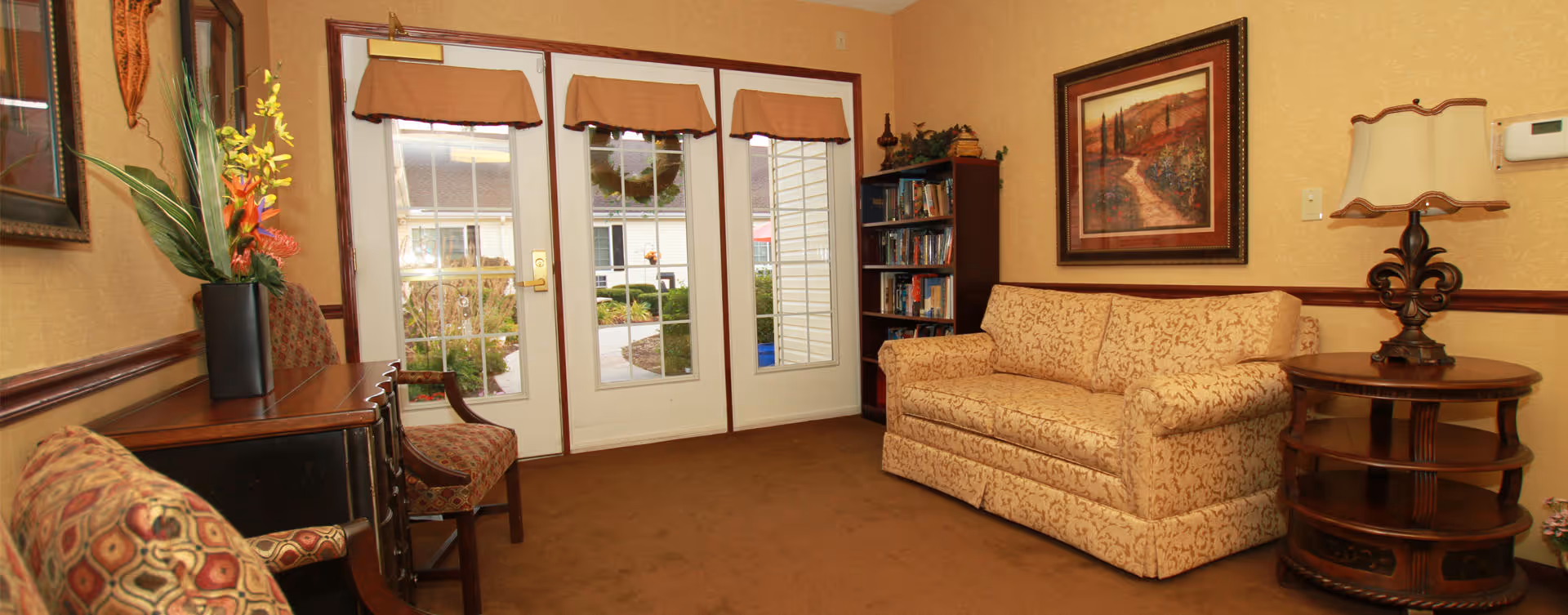 A cozy living room area with a patterned beige sofa, a wooden side table with a decorative lamp, a bookshelf filled with books, and two upholstered chairs. The room has beige walls with framed artwork and a glass door leading to an outdoor garden area.