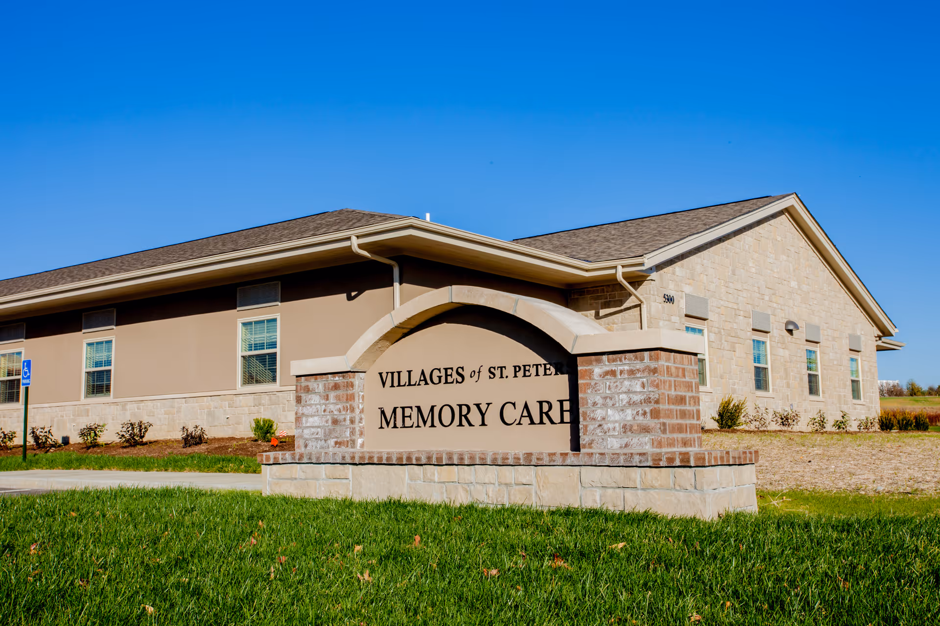 Exterior view of a single-story building with beige and stone walls under a clear blue sky. In front of the building is a large stone and brick sign that reads 'Villages of St. Peters Memory Care'. There is a small landscaped area with grass and shrubs around the sign and building.
