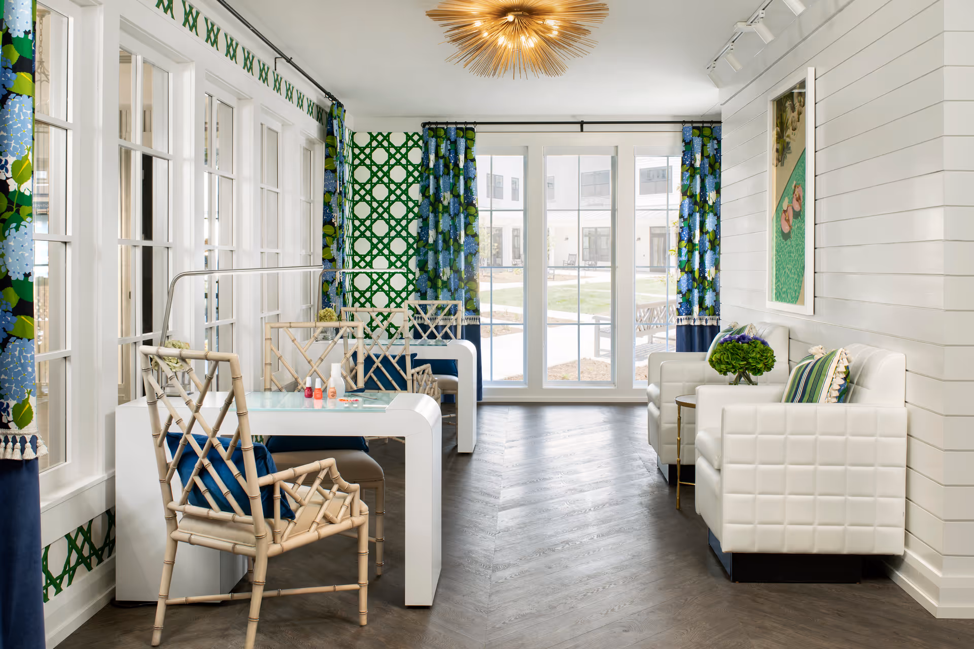 A bright and stylish interior room with large windows and glass doors leading outside. The room features white walls with green patterned wallpaper on one side, blue and green floral curtains, a white modern table with bamboo-style chairs, and two white cushioned armchairs with a small side table holding a green plant. A decorative light fixture hangs from the ceiling.