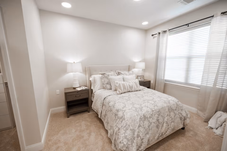 Neutral-toned bedroom with a made bed, two nightstands with lamps, and a window with blinds and curtains.