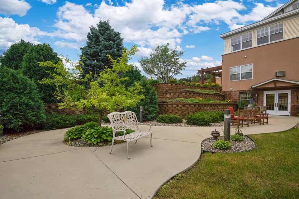Outdoor patio area at River Heights by Vivie featuring a curved concrete walkway, a decorative metal bench, wooden chairs and table, landscaped garden beds with green shrubs and trees, and a multi-level retaining wall with flowers under a partly cloudy sky.