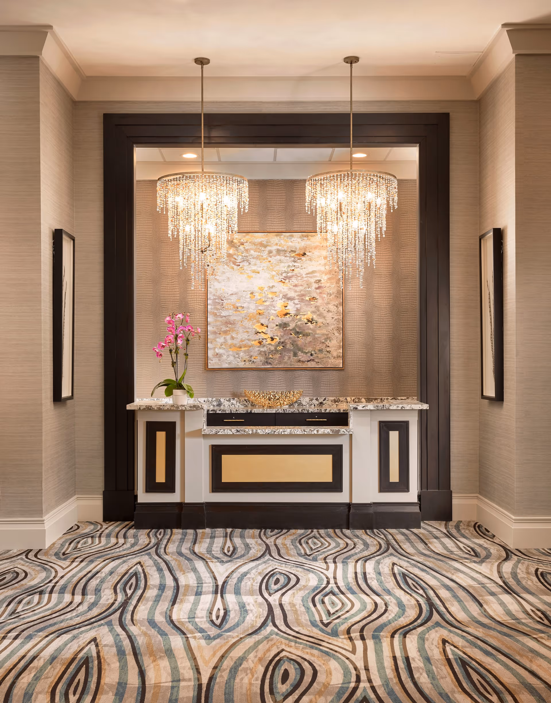 Elegant interior space featuring a decorative console table with a marble top, two crystal chandeliers hanging above, a large abstract painting on the wall, and a patterned carpet with swirling designs in shades of blue, beige, and brown. There are framed artworks on both side walls.