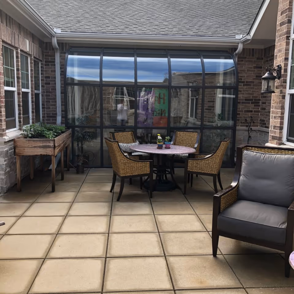 Outdoor patio area with a tiled floor, featuring a round table with four wicker chairs and a cushioned armchair. There is a wooden planter box with green plants on the left side and a glass-enclosed sunroom or greenhouse attached to a brick building in the background.