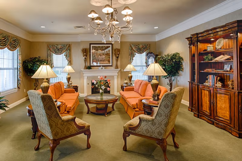 A warmly lit senior living facility common area with two orange sofas facing each other, two patterned armchairs, a round glass coffee table with a vase of flowers, several table lamps, a chandelier, large windows with decorative curtains, a fireplace with a framed picture above it, and a wooden bookshelf filled with books and decorative items.