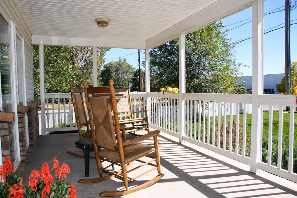 A covered porch with wooden rocking chairs and a bench overlooking a green lawn and trees under a clear blue sky.