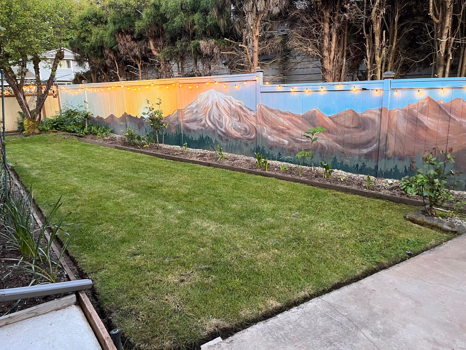 A backyard garden area with a well-maintained green lawn bordered by flower beds. A wooden fence painted with a mural of mountains and trees is decorated with string lights. Trees and shrubs are visible around the garden.