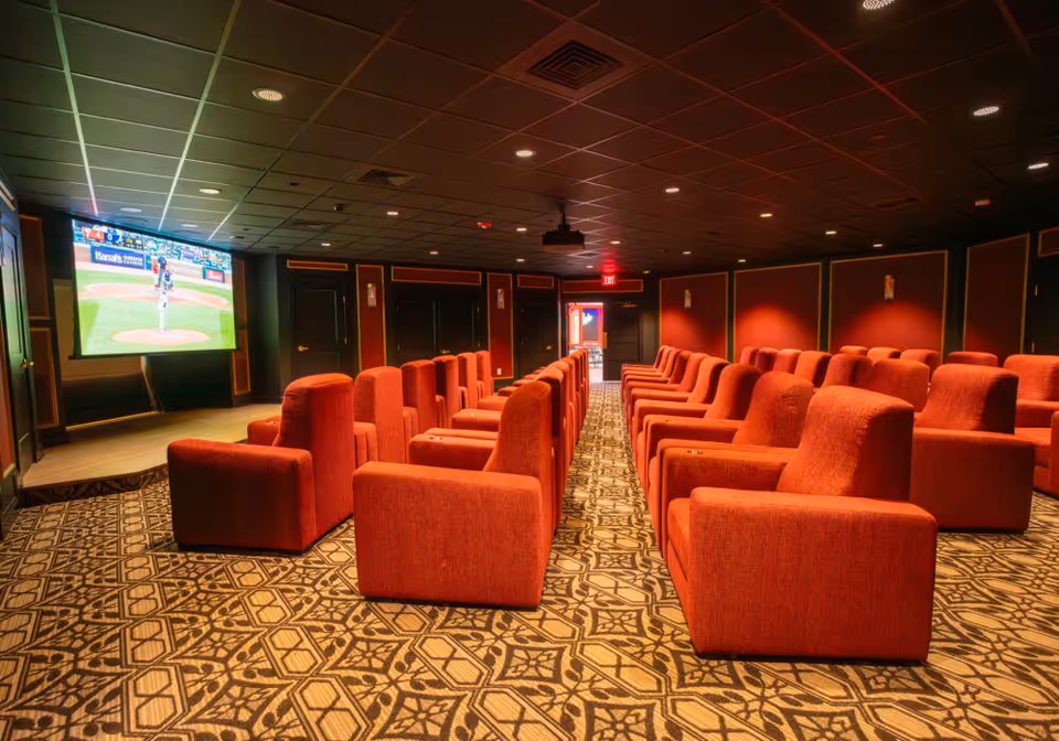 A cozy theater room with multiple rows of plush red armchairs facing a large screen displaying a baseball game. The room has patterned carpet flooring, dark walls with red accents, and a ceiling with recessed lighting.