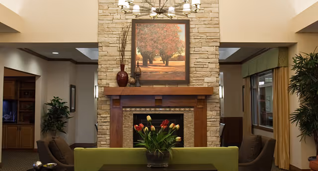 A cozy living room area featuring a stone fireplace with a wooden mantle. Above the fireplace hangs a framed painting of a tree-lined path with autumn foliage. In front of the fireplace is a green sofa with a vase of tulips on a dark coffee table. The room is warmly lit with a modern chandelier above and has comfortable armchairs on either side. There are plants and windows with curtains in the background.