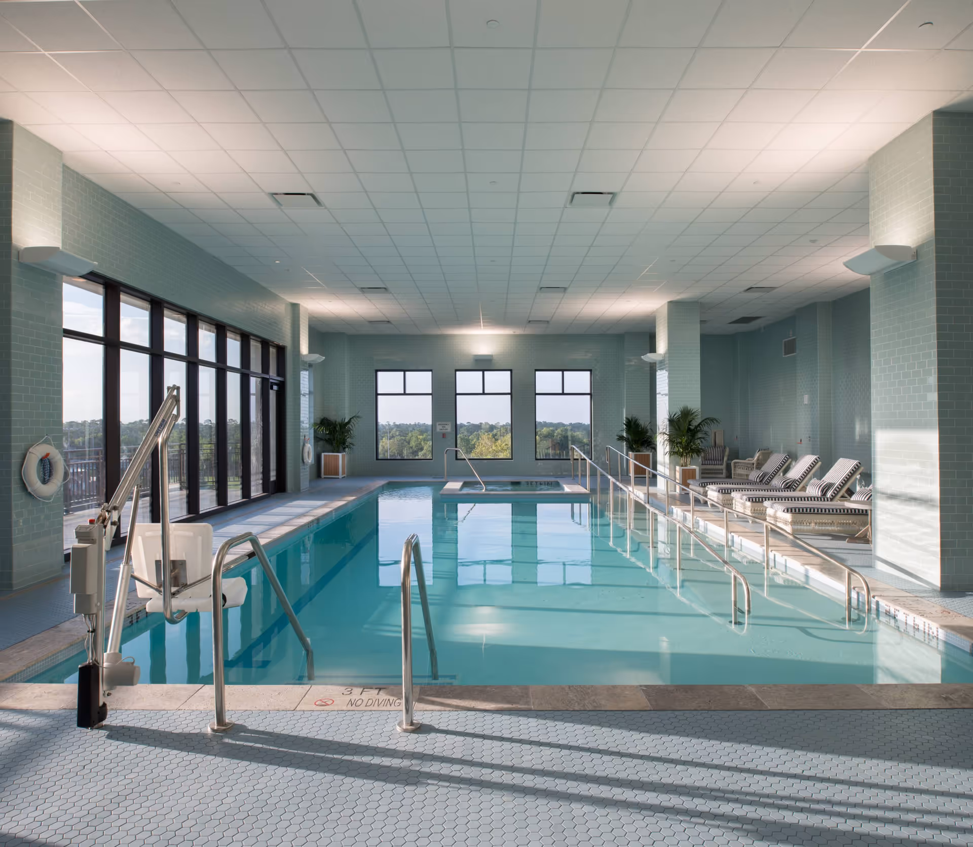 Bright indoor swimming pool with a lift chair, handrails, lounge chairs and large windows overlooking trees.