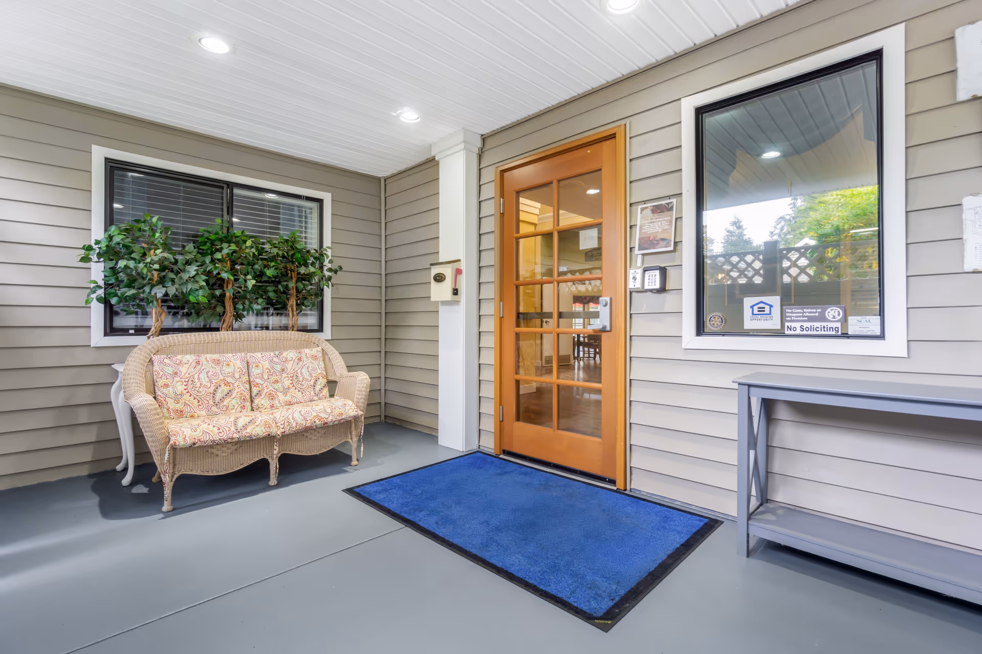 Covered entrance area with a wooden door featuring glass panels, a blue floor mat, a wicker loveseat with patterned cushions, potted plants behind the loveseat, and a gray table against the wall. There are two windows, one with several signs including 'No Soliciting'.