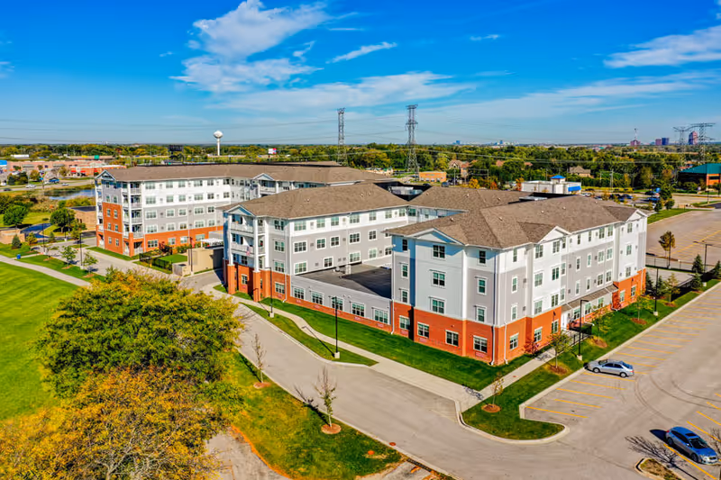 Aerial view of Clarendale of Addison, a multi-story senior living facility with red brick and white exterior walls, surrounded by green lawns, trees, parking lots, and nearby buildings under a blue sky with scattered clouds.