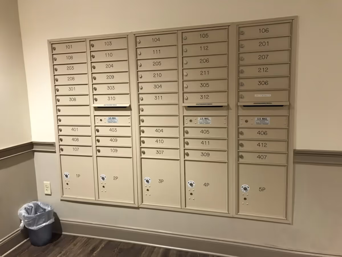 A set of beige mailboxes mounted on a wall inside a building, each labeled with different apartment or room numbers. There is a small trash can with a plastic liner on the floor to the left of the mailboxes. The wall is painted in two tones with a chair rail separating the colors, and the floor has a wood-like finish.