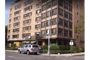Exterior view of a multi-story brick building with several windows, a covered entrance, and a parked silver SUV in front. There are street signs and a small tree near the sidewalk.