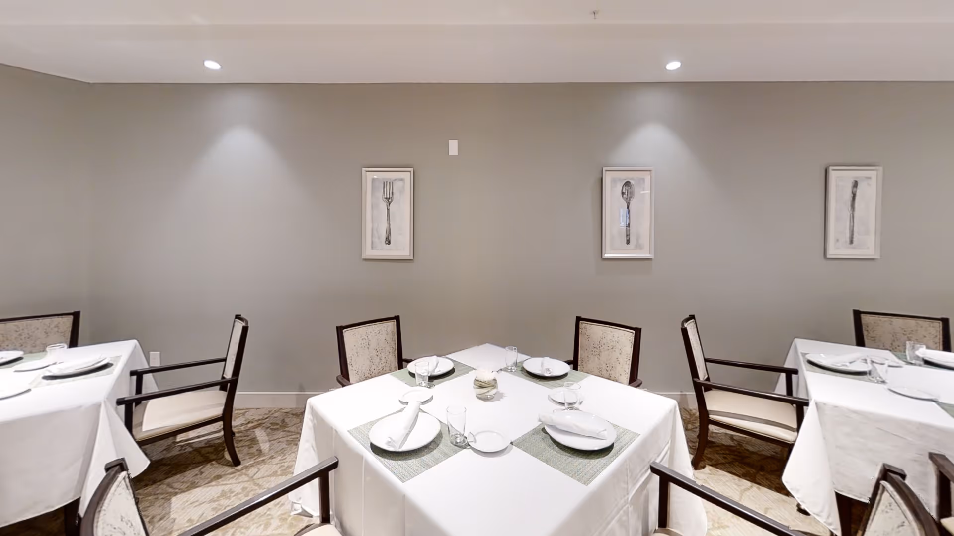 A dining room with three tables covered in white tablecloths, each set with plates, glasses, and napkins. The chairs have wooden frames with cushioned seats and backs. The walls are painted light gray and decorated with framed pictures of utensils. The room is well-lit with recessed ceiling lights.