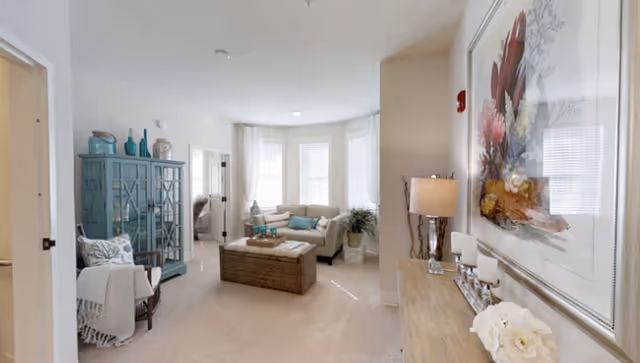 A bright and cozy living room with beige carpet and walls. The room features a beige sectional sofa with teal and beige cushions, a wicker coffee table, and a teal cabinet with decorative items on top. A wooden console table with a lamp, candles, and a large floral painting is on the right side. Large windows with white curtains allow natural light to fill the space.