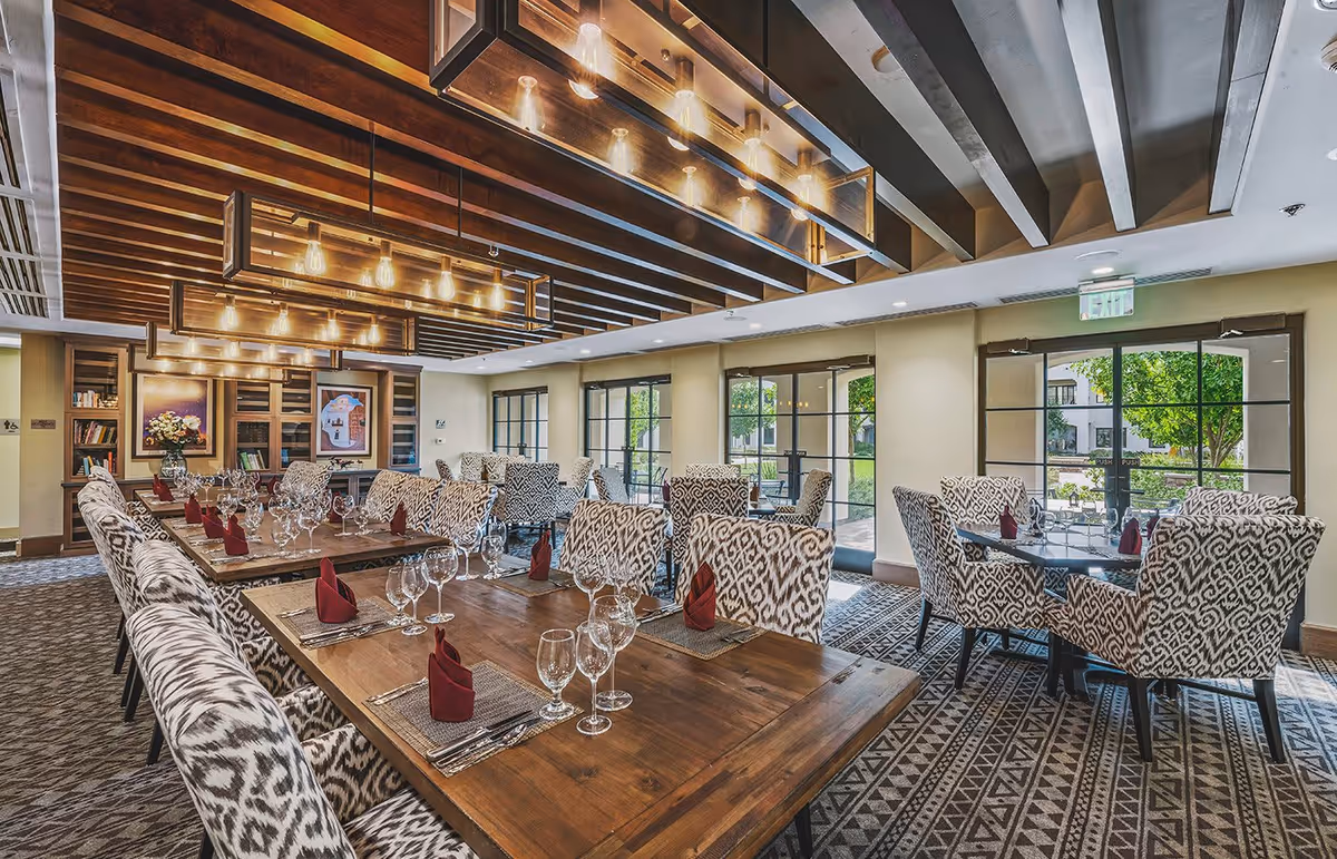 A well-lit dining room with wooden tables set with wine glasses, silverware, and folded red napkins. The chairs have a patterned upholstery, and large windows allow natural light to enter, showing greenery outside. The ceiling features wooden beams and modern hanging light fixtures.