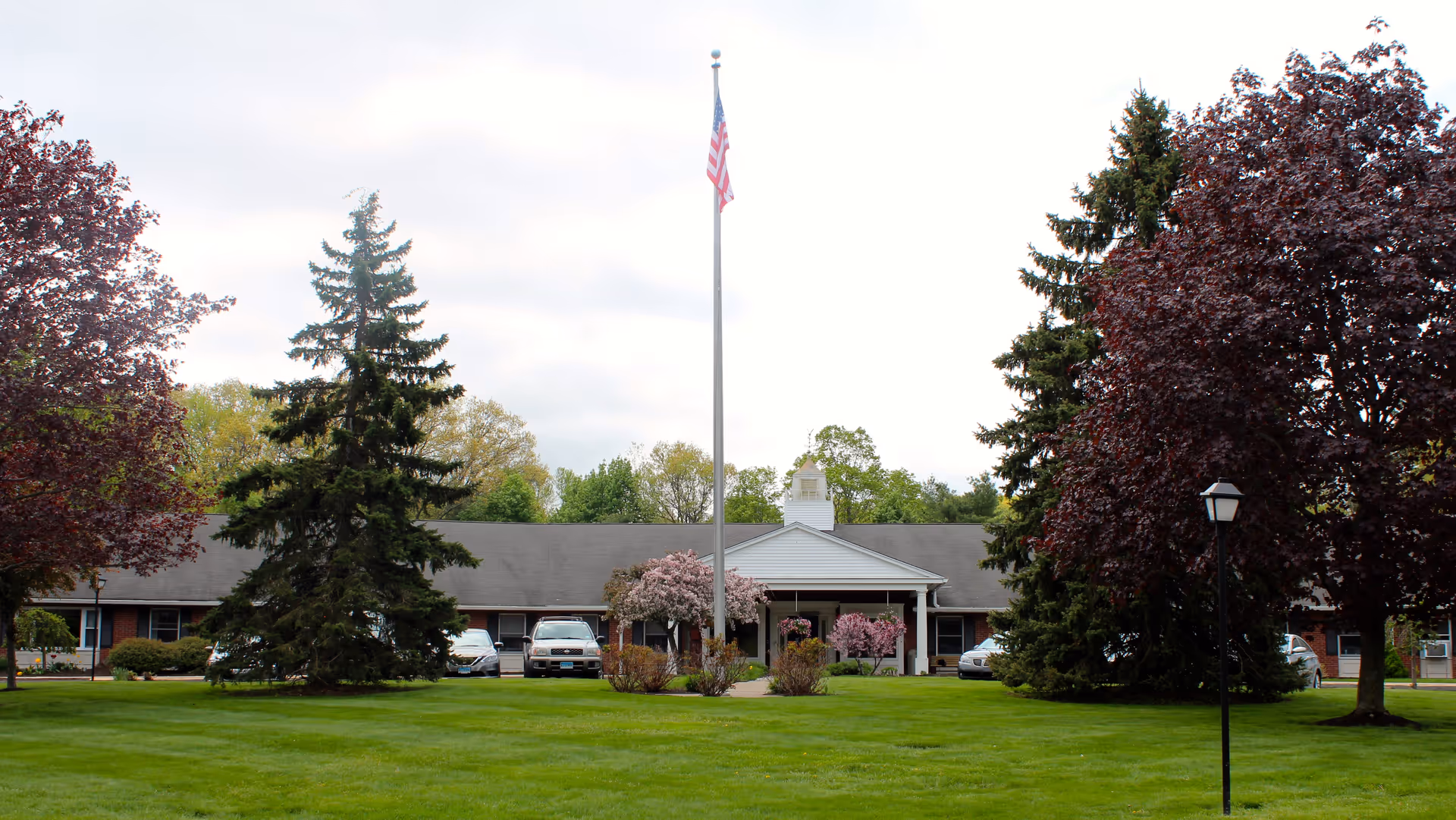 Front exterior view of a single-story brick building with a gray roof, surrounded by green grass, trees, and shrubs. An American flag is flying on a flagpole in the center of the lawn. Several cars are parked near the building entrance.