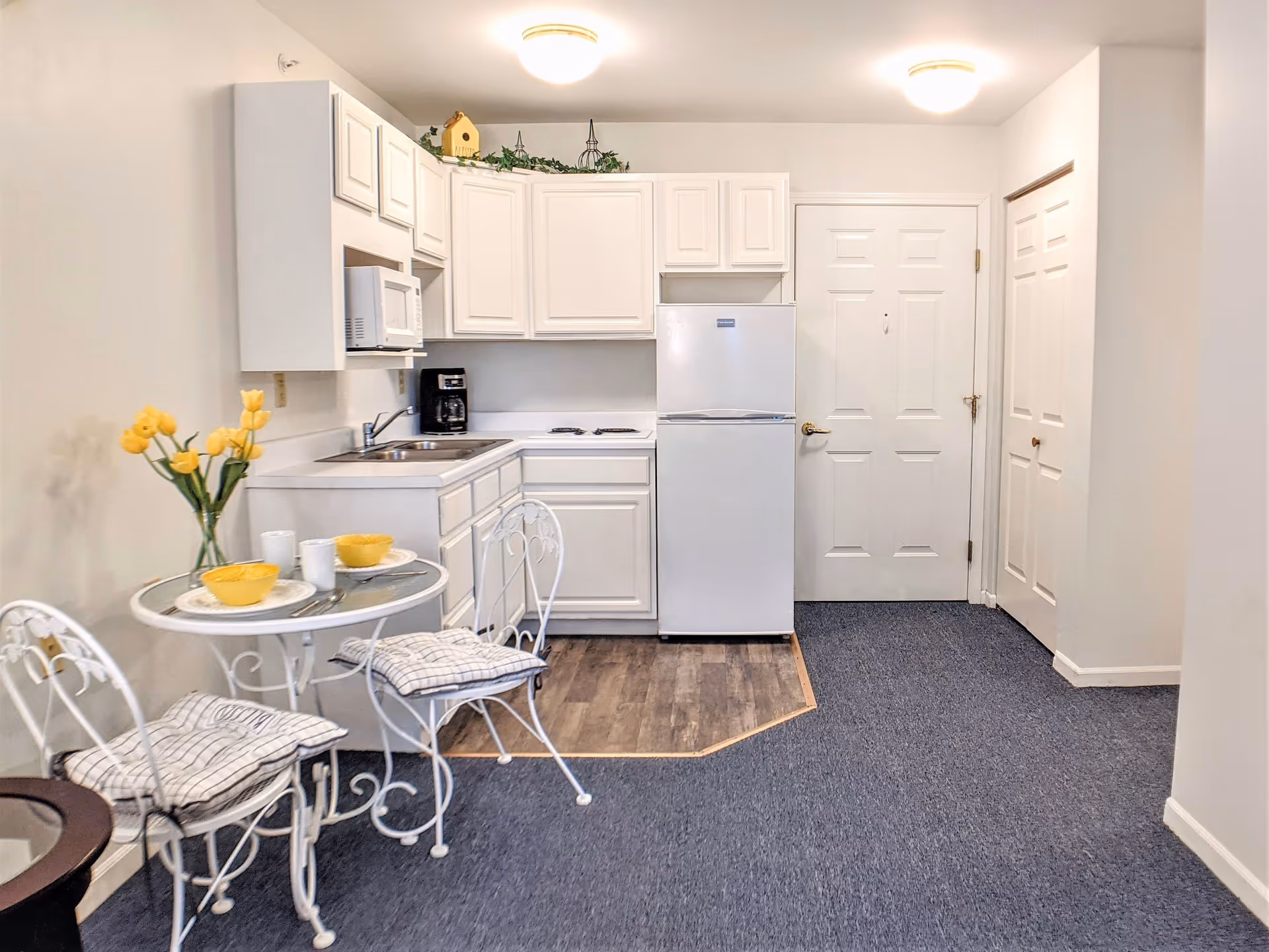 Compact kitchenette with white cabinets and appliances, a small round bistro table with two chairs, and a door leading to another room.