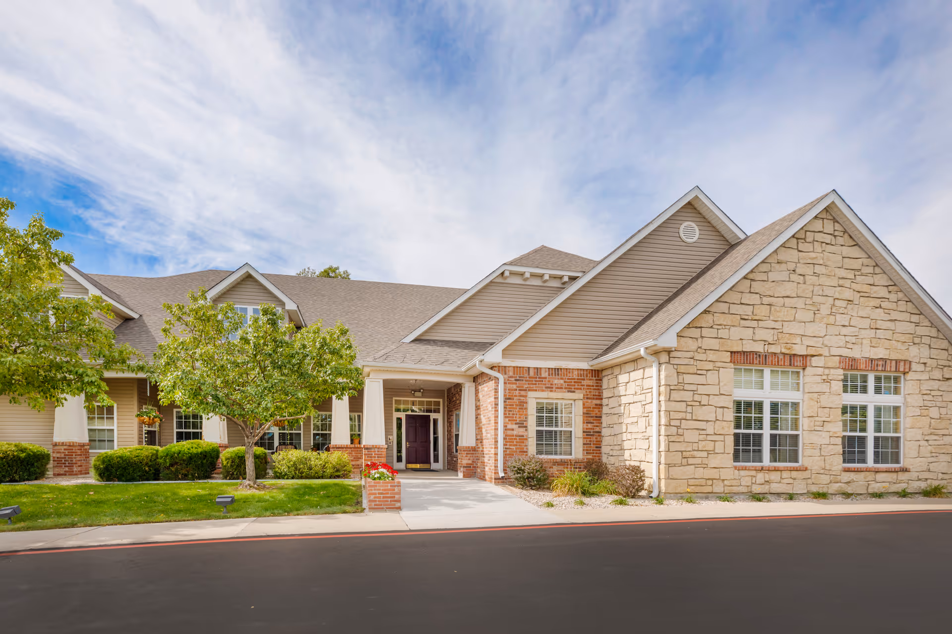 Exterior view of a single-story senior living facility building with a combination of stone and brick facade, a covered entrance with white columns, green bushes, trees, and a clear sky with some clouds.