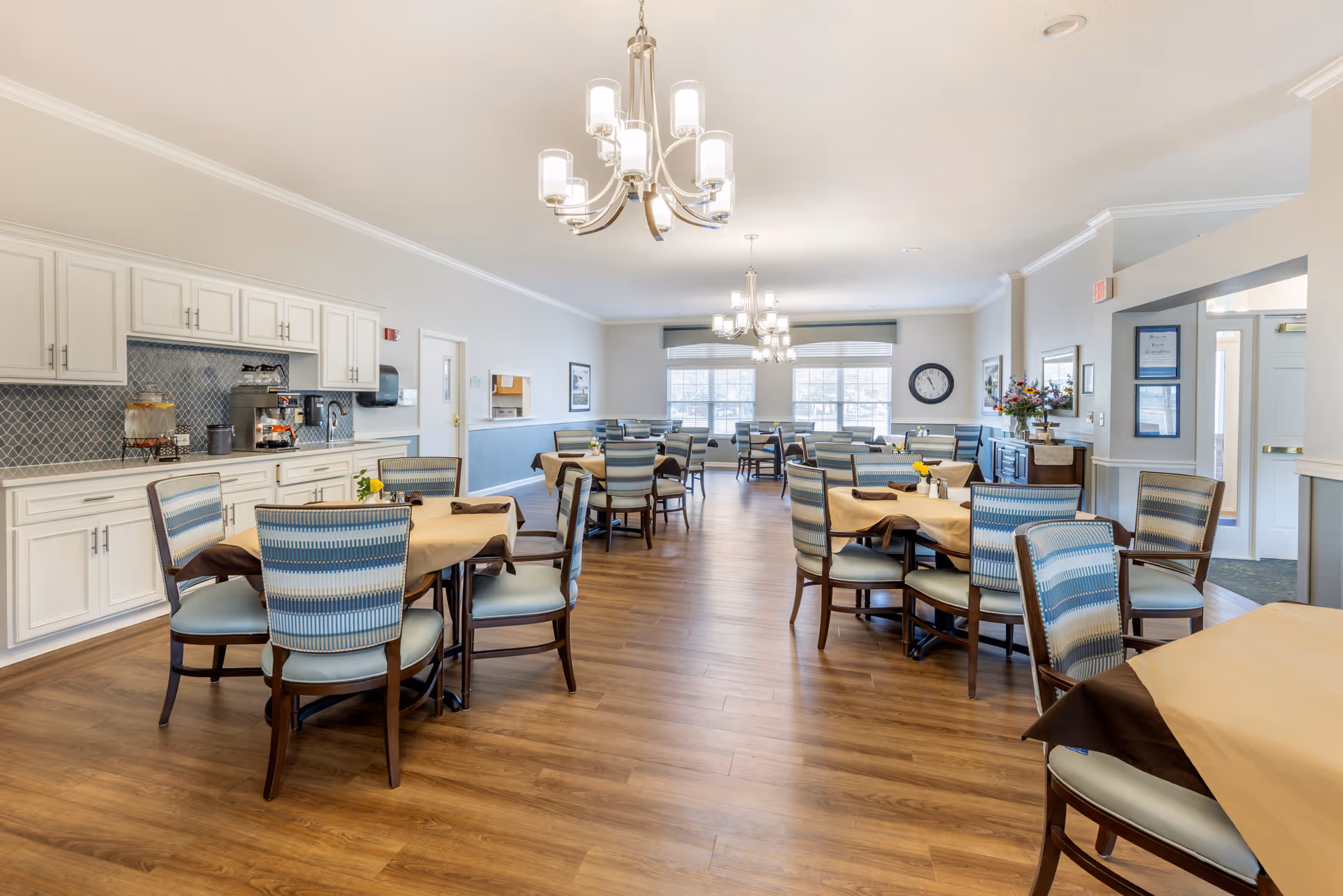 A spacious dining room with multiple tables covered in beige tablecloths and surrounded by chairs with blue and white striped upholstery. The room features wooden flooring, white cabinetry with a coffee and beverage station on the left, large windows letting in natural light, and modern chandeliers hanging from the ceiling. There are decorative elements such as a wall clock, framed pictures, and a flower arrangement on a sideboard.