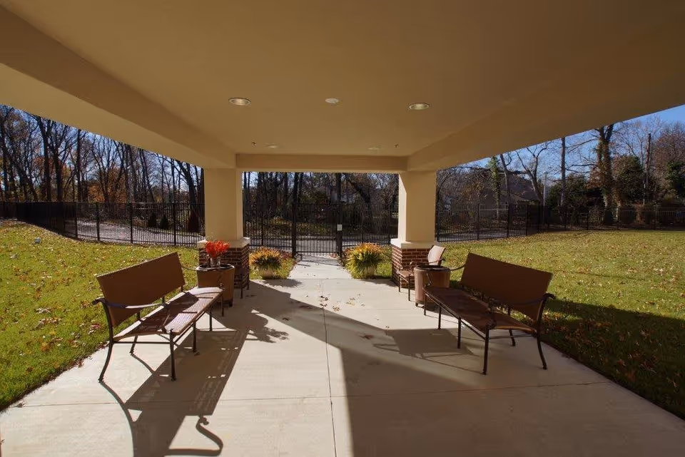 Covered outdoor seating area with two brown benches facing each other, small tables with potted plants, and a view of a fenced grassy yard with trees in the background under a clear blue sky.