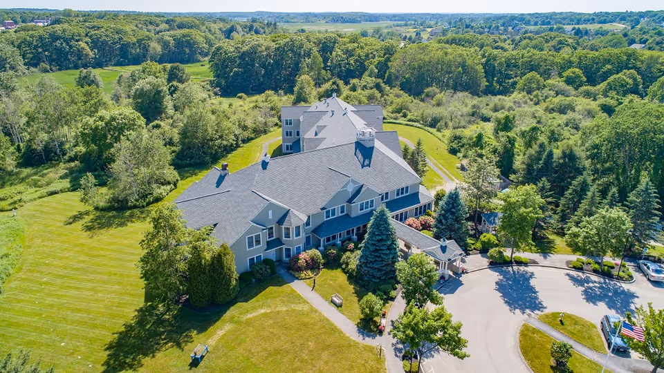 Aerial view of a large senior living facility building surrounded by green lawns, trees, and landscaped gardens under a clear blue sky.