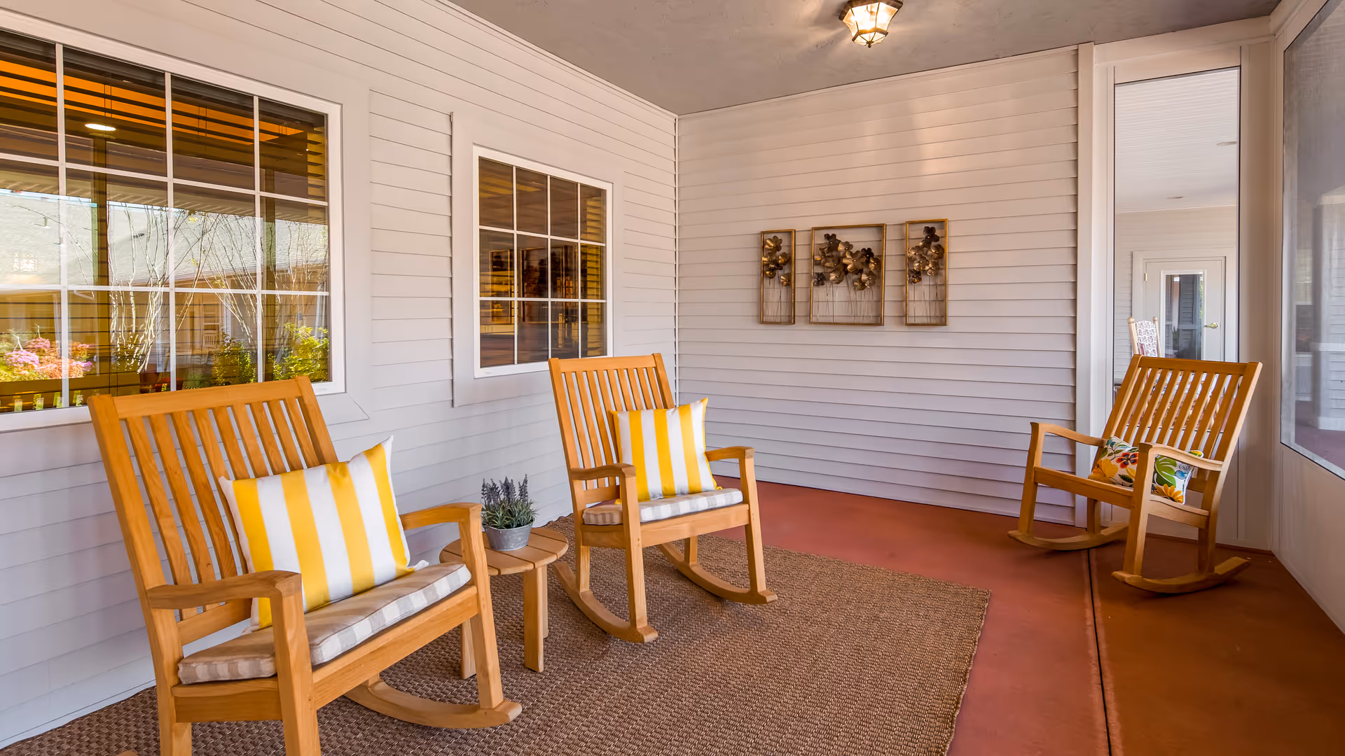 A cozy covered porch area with three wooden rocking chairs, two with yellow and white striped cushions and one with a floral cushion. There is a small round wooden table with a potted plant between two of the chairs. The porch has white siding walls, two windows, a ceiling light fixture, and decorative wall art featuring metal flowers. The floor is covered with a brown rug and reddish-brown concrete.