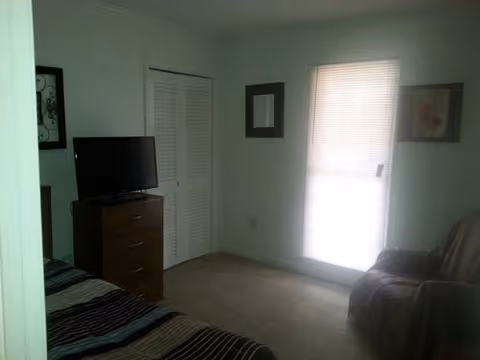 A small bedroom with a bed partially visible on the left, a wooden dresser with a flat-screen TV on top, a closed white closet door, a window with blinds letting in light, and a cushioned chair on the right side.
