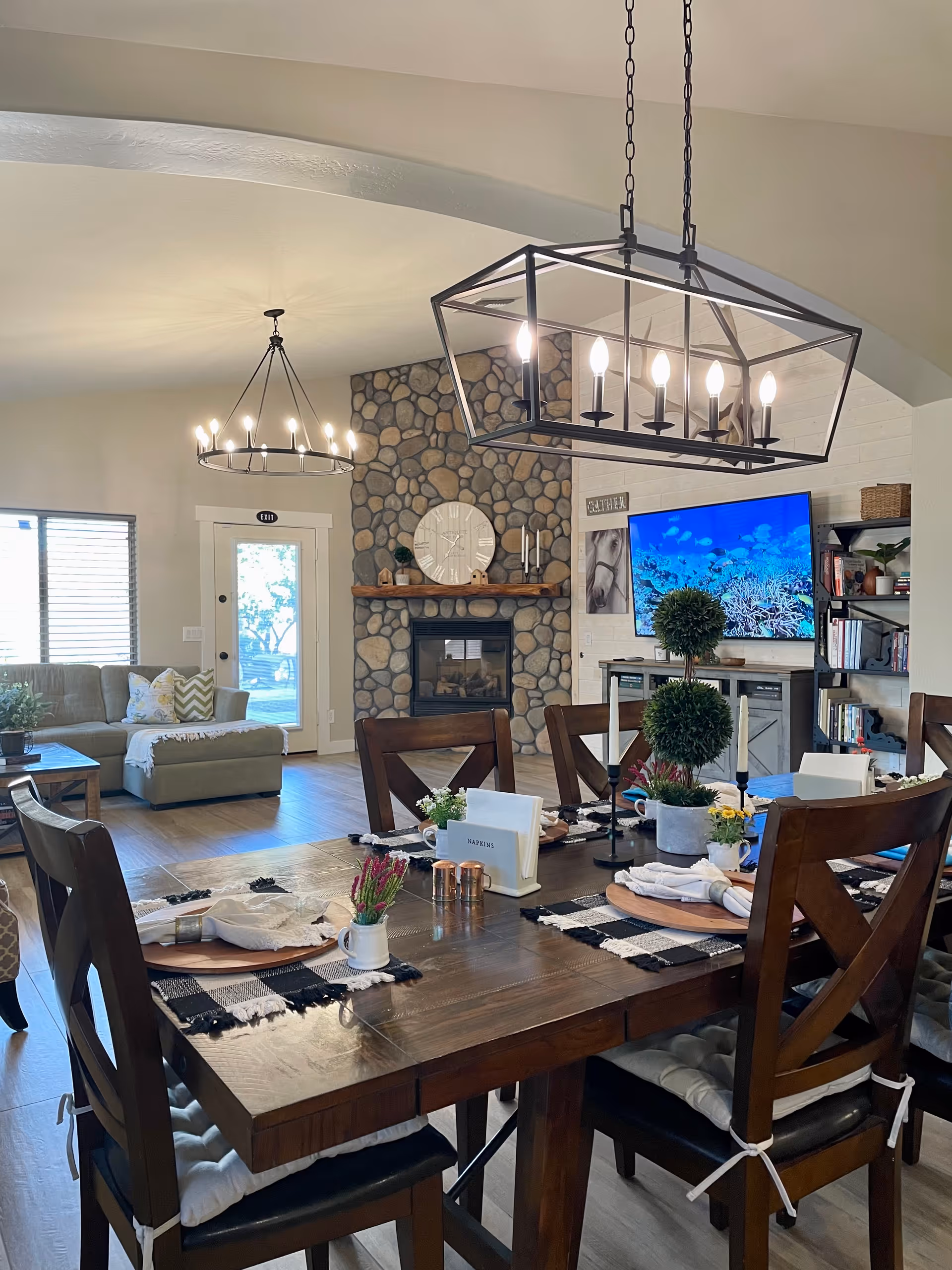 Open-plan dining and living area with a wooden dining table set for place settings, chandeliers overhead, a stone fireplace, sofa, and wall-mounted TV.